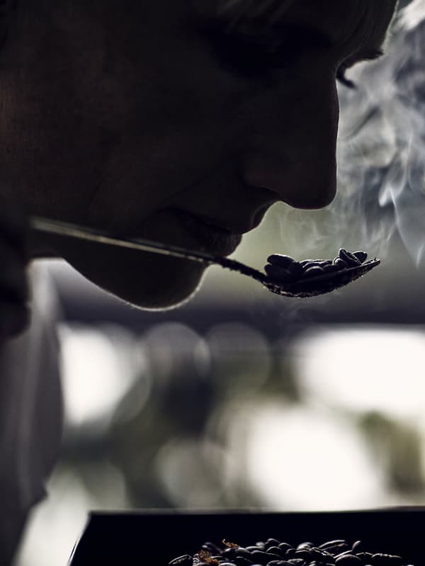 A silhouette of a person smelling roasted coffee beans on a spoon. Steam is visible coming off the spoon.