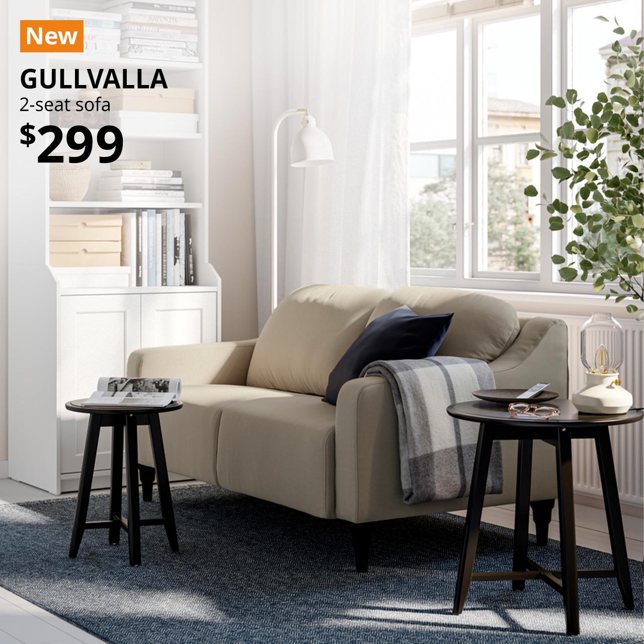 A silkeryd dark grey-beige GULLVALLA two-seater sofa placed against the living room window next to a white HAUGA high cabinet. The cabinet holds a projector, books, and small storage organisers, showcasing a functional and organised home storage solution. An orange "NEW" label is displayed in the top left corner of the image, featuring white text that reads: "GULLVALLA, 2-seat sofa, $299".
