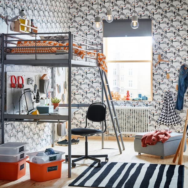 A silver steel loftbed with a sturdy desk on the bottom bunk in a room with black and white patterned wallpaper.