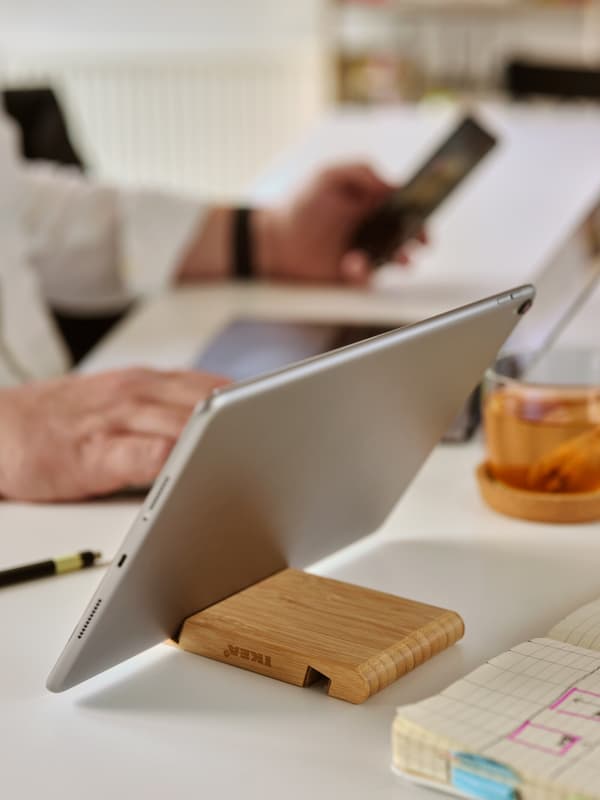 A silver tablet in a bamboo BERGENES holder on a white desk, in front of a person behind a laptop with a smartphone.