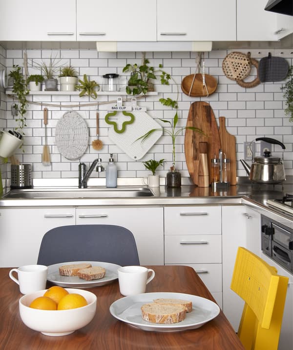 A simple breakfast laid out on a wooden table in a small white kitchen with plants and utensils hung on the walls.
