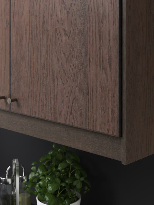 A SINARP cabinet door in dark wood with NYDALA knobs in bronze. There is a pot of basil and condiments underneath.