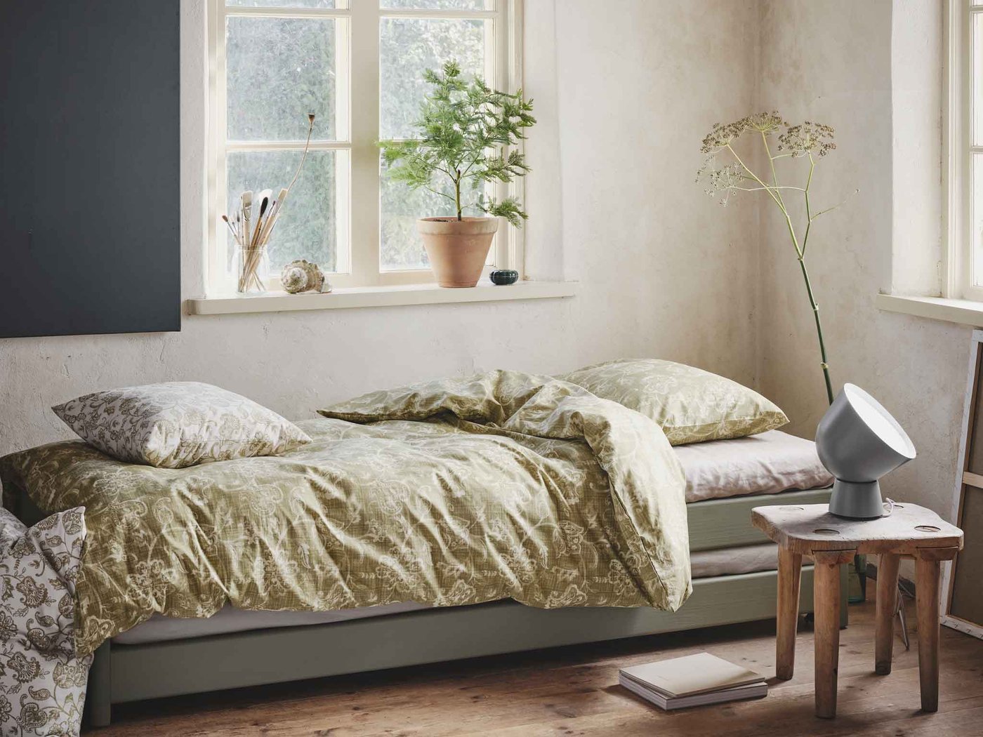 A single bed in the corner of a room with a green-yellow patterned duvet on top with matching pillows and a lamp next to it