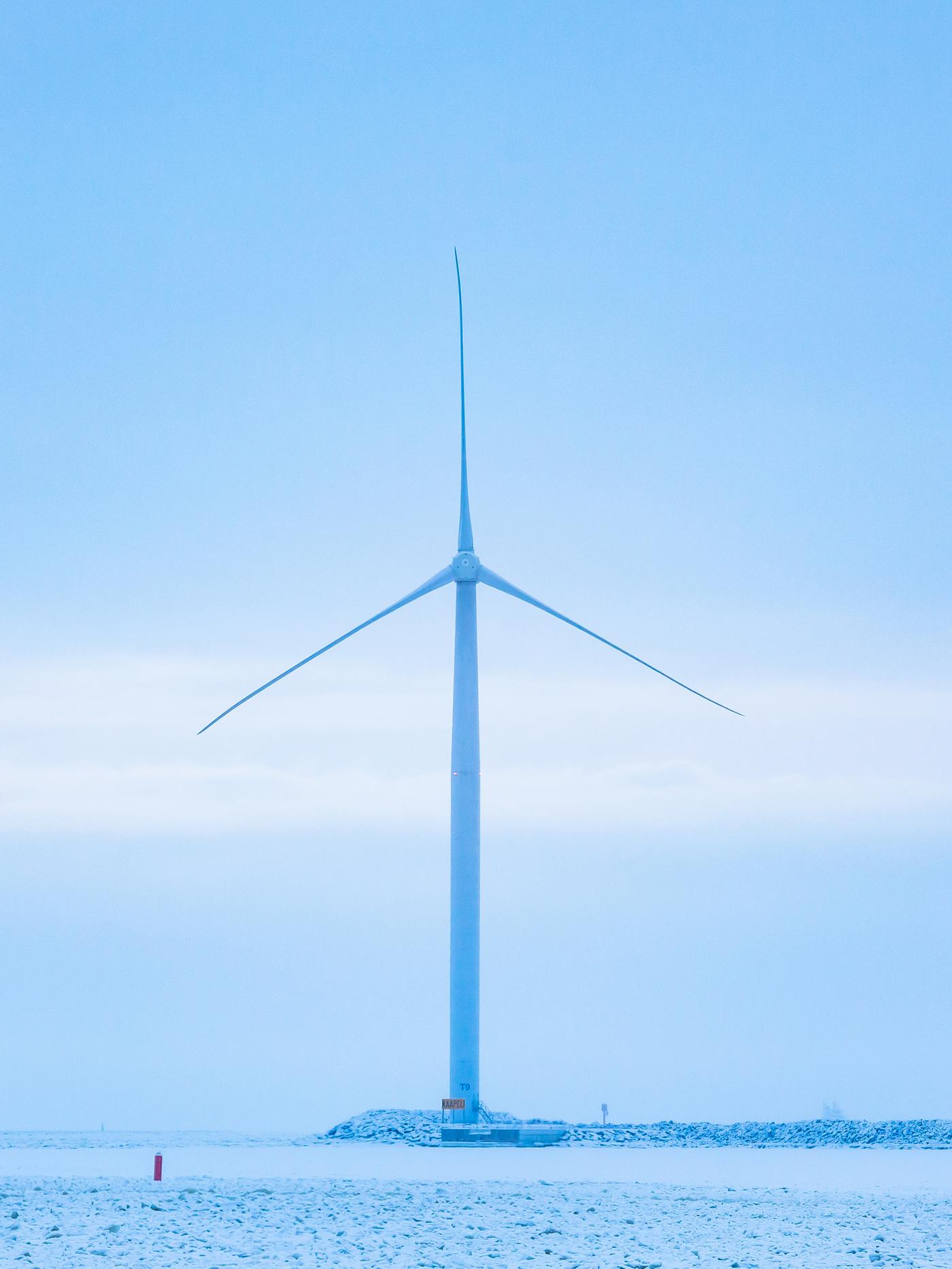 A single wind turbine at the end of a stone breakwater. The sky is grey blue with a single band of light grey cloud.