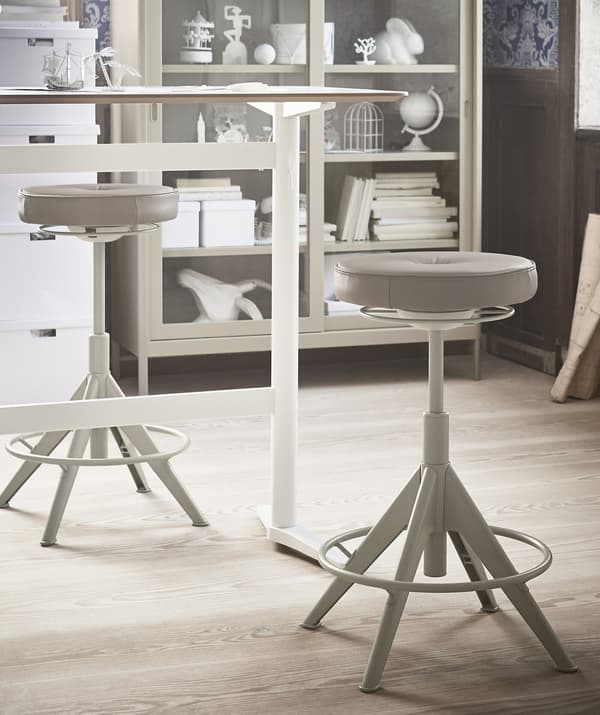 A sit/stand desk with two beige stools and a display cabinet in the background.