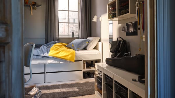 A SLÄKT bed with blue bed linen in front of a window in a small bedroom with TROFAST storage units on the wall and floor.