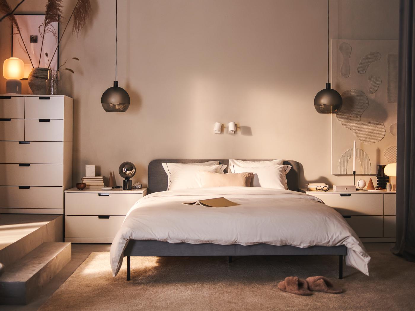 A SLATTUM upholstered bed and various white NORDLI chests of drawers stand against a wall in a bedroom.