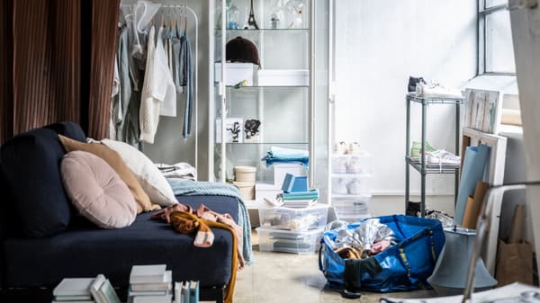 A slightly messy living room with a blue sofa, a shoe rack, some stackable storage boxes and a half-full FRAKTA bag.