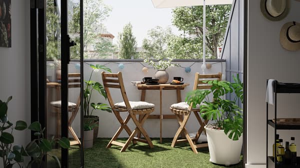 A small balcony with a wooden table and chairs, artificial grass decking, and MONSTERA plant in a white plant pot.