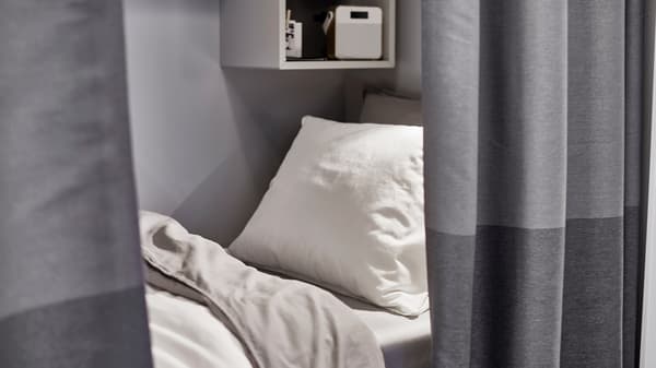 A small bed with a white pillow and light-grey bed linen behind a dark-grey curtain room divider.