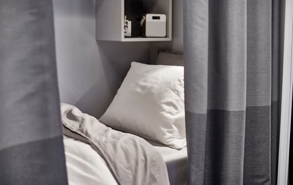 A small bed with a white pillow and light grey bedlinens peeking out from behind a dark grey curtain room divider.