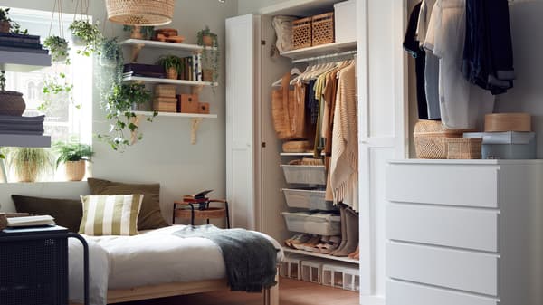 A small bedroom with a wooden bed frame, a wardrobe system with neatly organised clothes, shelves with boxes and plants.