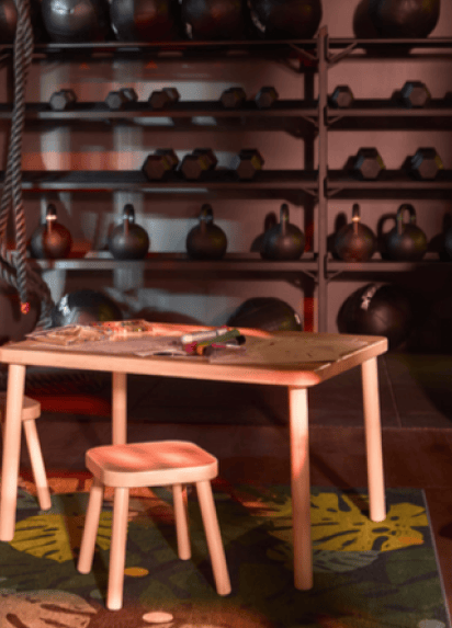 A small birch table with a stool on a rug with green leaves, work out gear in the background on grey wall shelves.