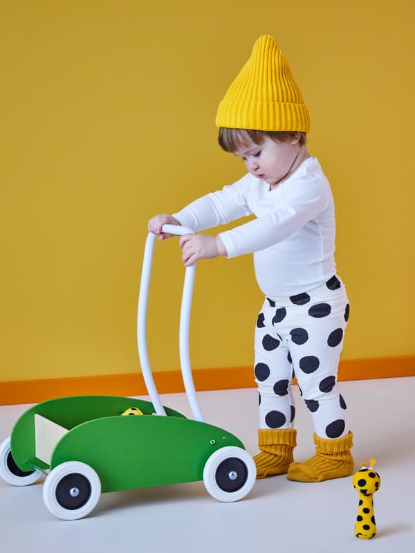 A small boy in a yellow woolly hat pushing MULA green child’s trolley with a KLAPPA small soft toy and a yellow wall.