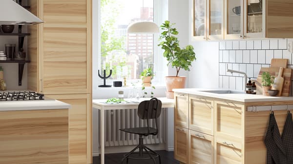 A small, bright kitchen with traditional natural ash and glass cabinets and a white desk in front of a window.
