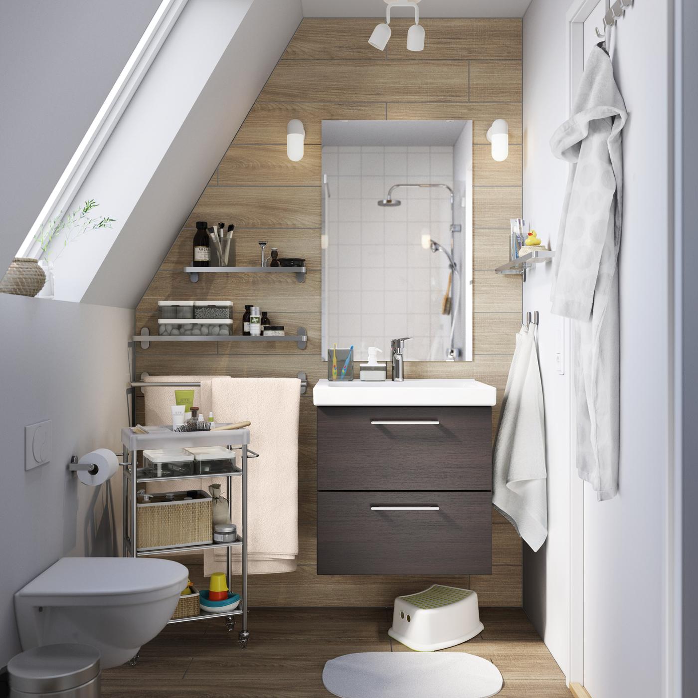 A small brown and white bathroom with a sloping ceiling and brown sink cabinet with drawers.