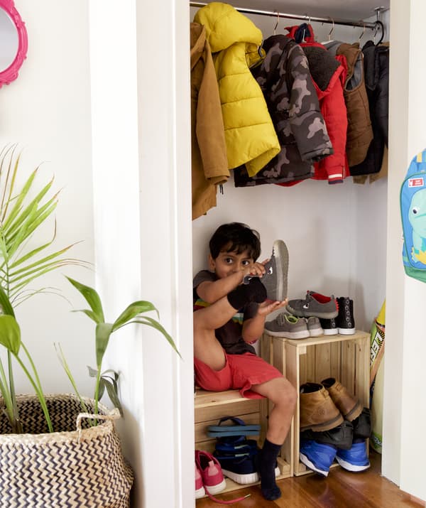 A small child putting shoes on and sitting on a wooden crate below coats hanging in a cupboard.