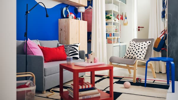 A small colourful living room has bold blue walls, a light grey FRIDHULT sofa-bed and a red TINGBY side table with castors.