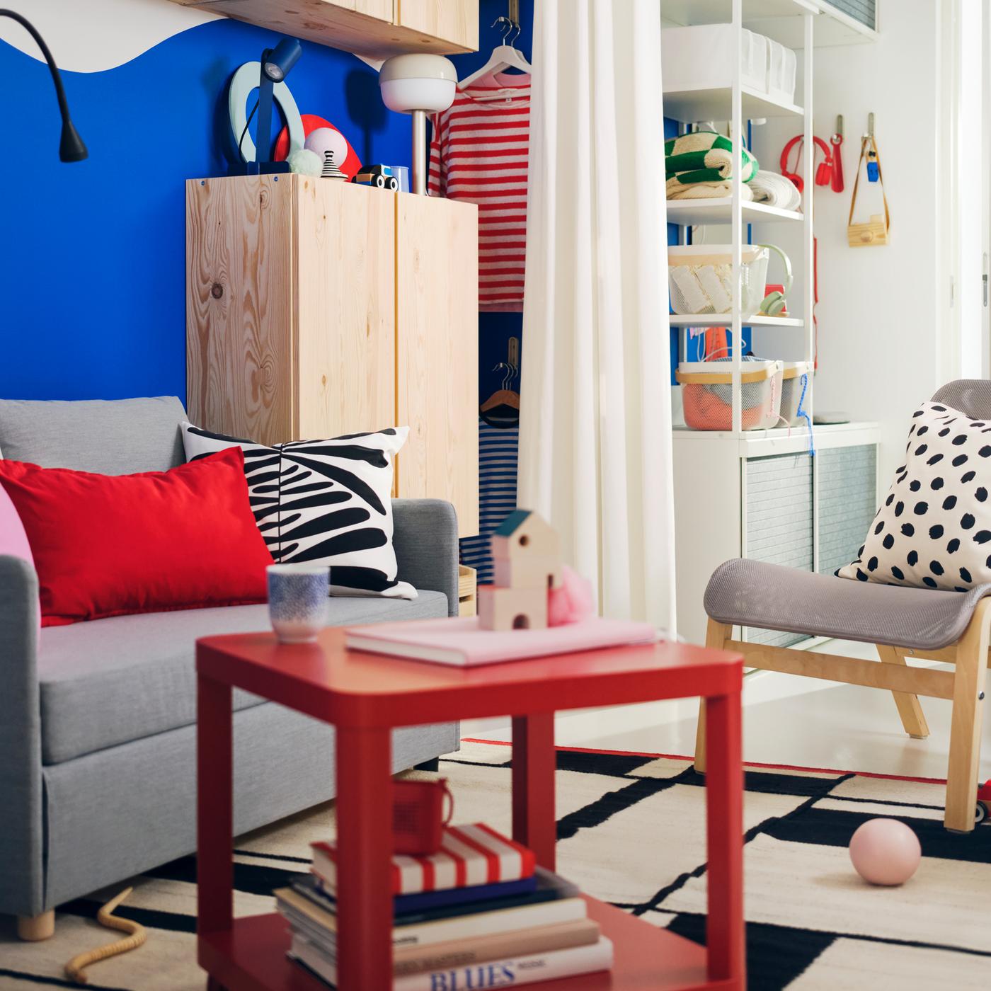 A small colourful living room has bold blue walls, a light grey FIRDHULT sofa bed and a red TINGBY side table with castors.