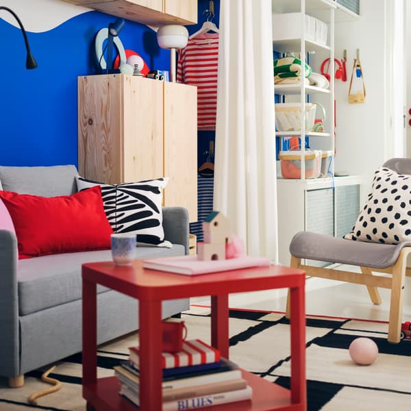 A small colourful living room has bold blue walls, a light grey FIRDHULT sofa bed and a red TINGBY side table with castors.