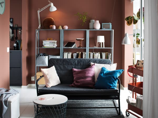 A small dark grey-black HAMMARN sofa in a living room setting with dark red walls and shelves across the back wall.