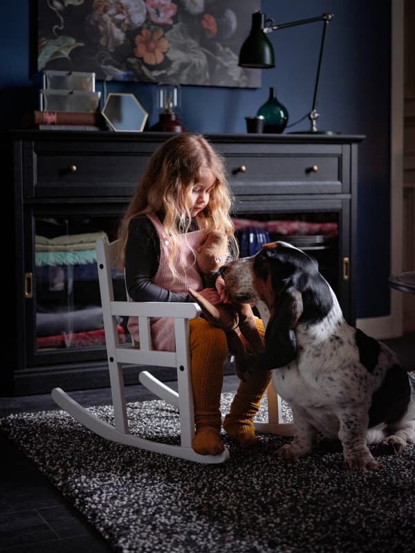 A small girl sits on a SUNDVIK rocking chair in a dark living room, while a basset hound rests its head in her lap.