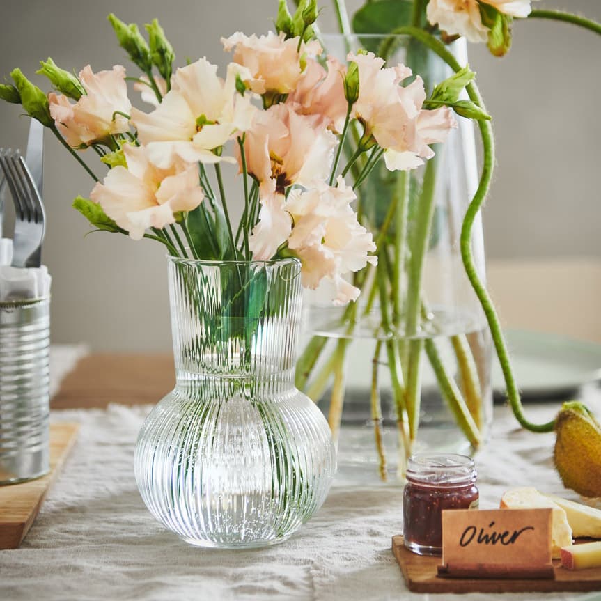 A small glass vase filled  with spring flowers sits on a traditionally decorated table. A cheese board with a name tag can be seen also. 