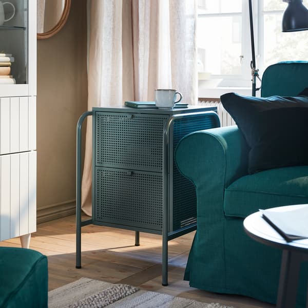 A small, grey-green chest of drawers with perforated surfaces, a dark turquoise sofa, a striped rug and a beige curtain.