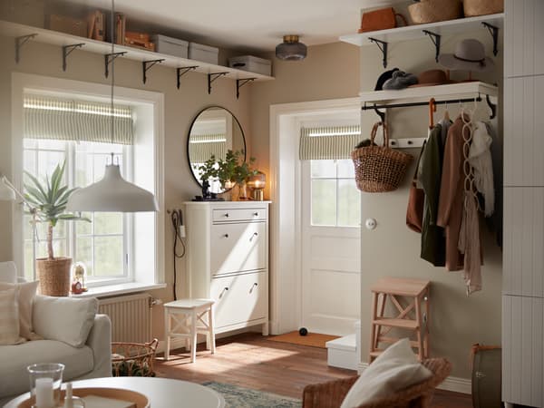 A small hallway with a white door, a white shoe cabinet, a round mirror, a white hat rack with jackets and an umbrella.