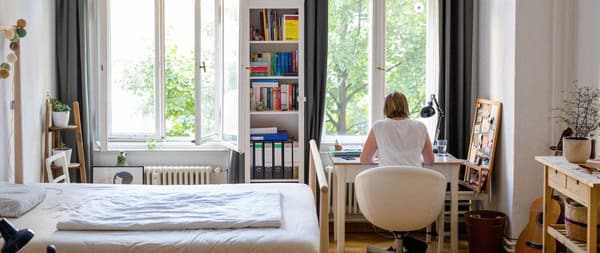 A small, light bedroom with a bookshelf and a desk with a young woman sitting with her back towards the camera.