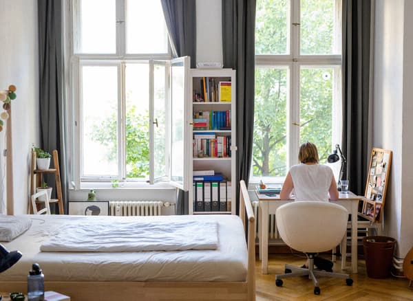 A small, light bedroom with a bookshelf and a desk with a young woman sitting with her back towards the camera.