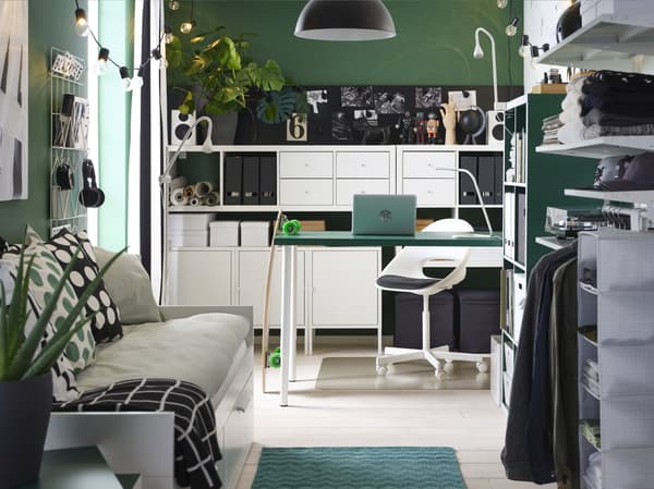 A small, overall green room with a green table, white shelving units, a day-bed, an open wardrobe and a black pendant lamp.