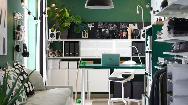 A small room with a green table, white shelving units, a day-bed, an open wardrobe and a black pendant lamp.