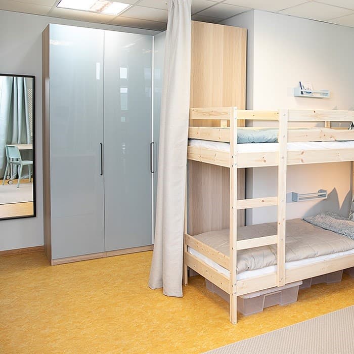 A small, tidy bedroom with a wooden bunk bed against the wall, a tall grey wardrobe beside it and a wall-mounted mirror reflecting part of the room.
