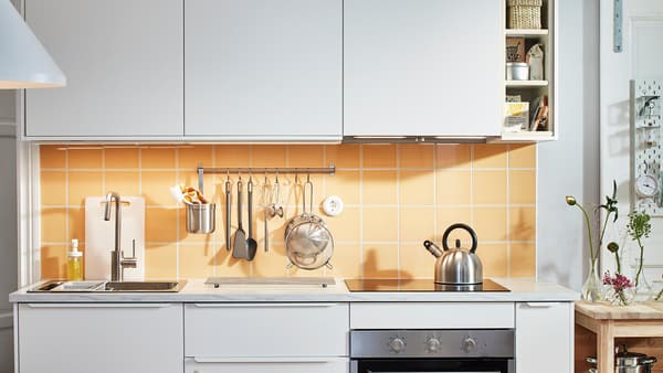 A small white kitchen with white VEDDINGE cabinet fronts and orange-yellow tiles, a hob, and a rail holding utensils.