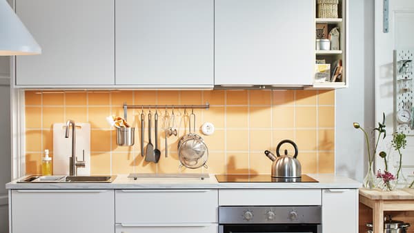 A small white kitchen with white VEDDINGE cupboard fronts and tiles in orange and yellow, a hob and a rail with utensils.