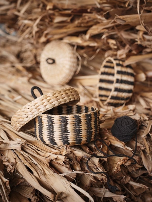 A small woven MÄVINN box with a lid, featuring a black stripe pattern, placed on a pile of natural banana fibre.