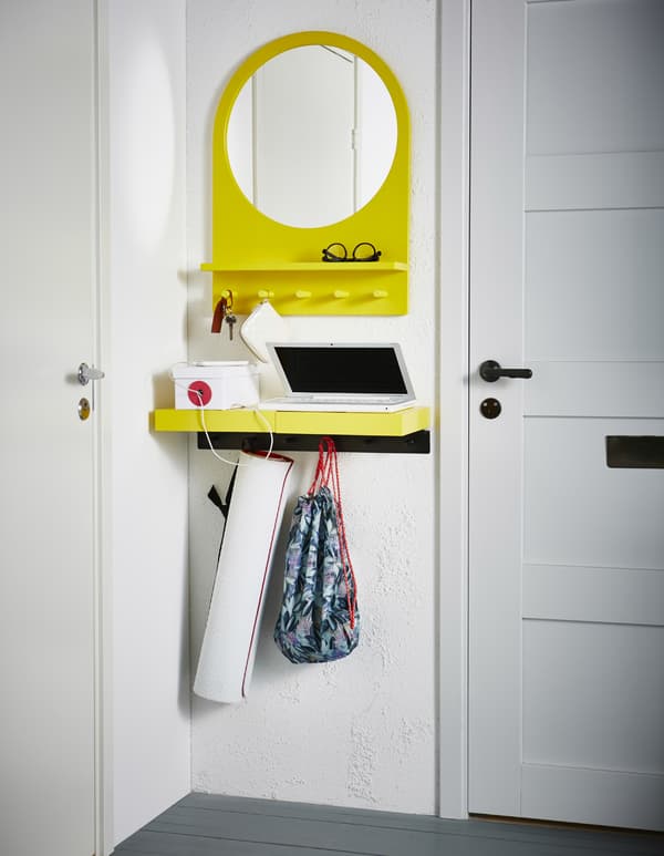 A small yellow wall shelf, hooks and mirror hang between a door fram and a corner, storing a laptop, phone, workout bag, yoga mat and glasses.