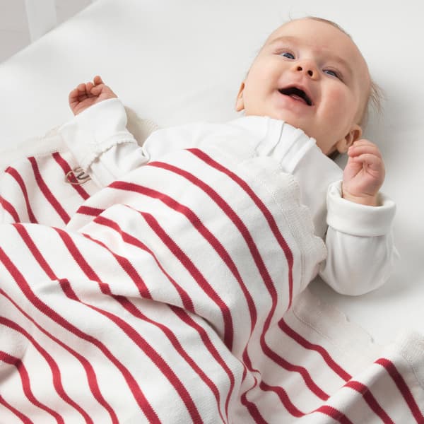 A smiling baby with a white and red striped RÖDHAKE blanket made from responsibly grown cotton.