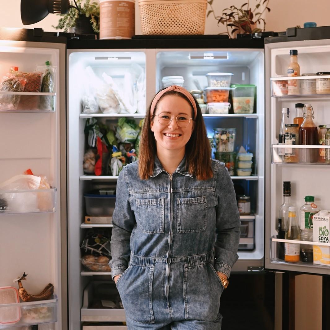 A smiling brunette in a denim jumpsuit stands in front of a refrigerator filled with various food items.