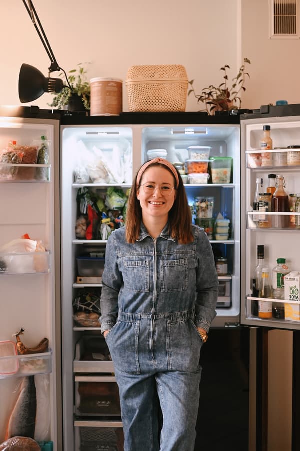 A smiling brunette in a denim jumpsuit stands in front of a refrigerator filled with various food items.				