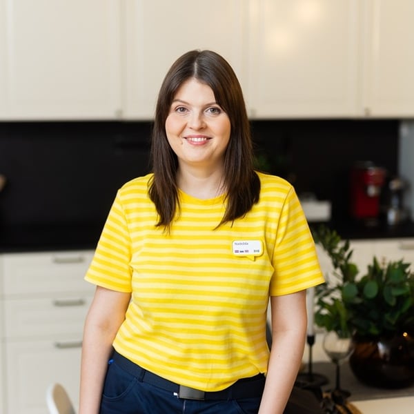A smiling IKEA co-worker standing in a kitchen, wearing a yellow striped shirt, with light-colored cabinets and plants visible in the background.