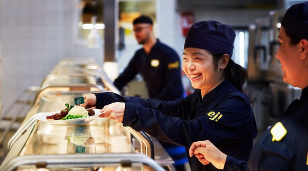 A smiling IKEA co-worker working in the IKEA restaurant, serving a plate of meatballs.