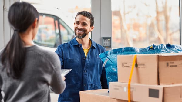A smiling IKEA delivery worker in a blue uniform is handing over a package to a customer at the entrance of a building, surrounded by cardboard boxes and a blue IKEA bag.