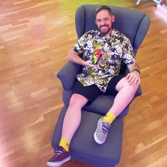 A smiling man is leaning back in a STRANDMON armchair. He has a video game console in his hand. The walls around him are covered with open shelves full of video games and toys.