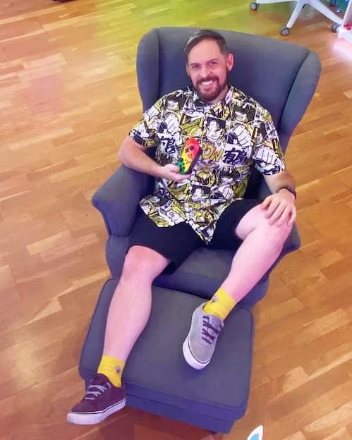 A smiling man is leaning back in a STRANDMON armchair. He has a video game console in his hand. The walls around him are covered with open shelves full of videogames and toys.