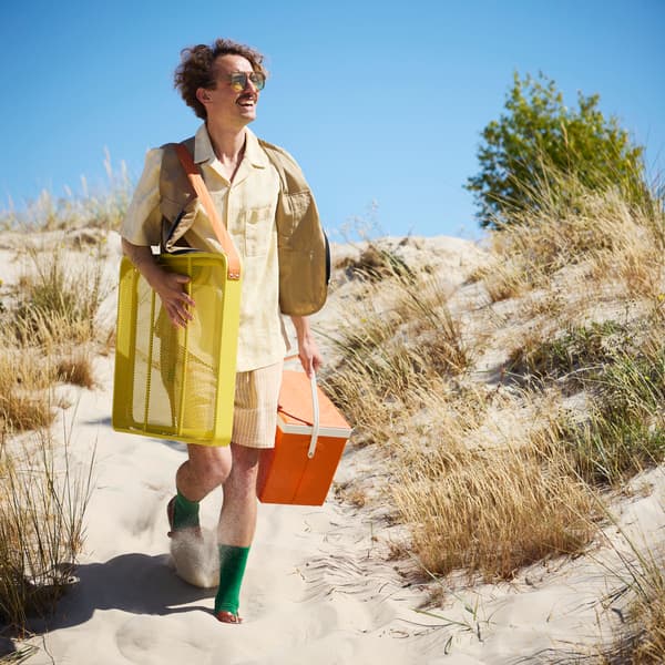 A smiling man walking on a sandy path carries a bright yellow STRANDÖN folding table and an orange cooling bag.