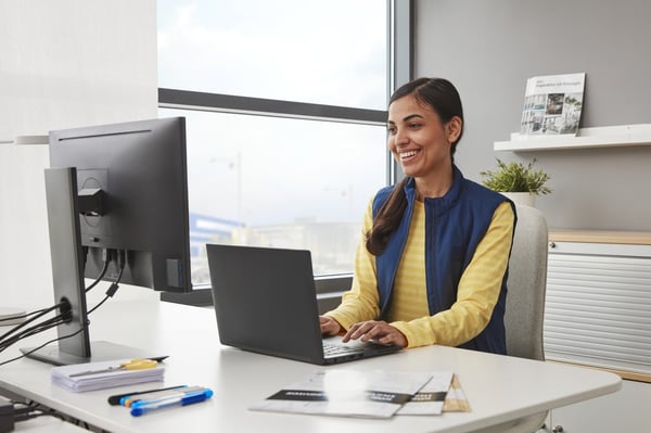 A smiling person in a yellow sweater and a blue vest is working on a black laptop placed on a white desk next to a window.