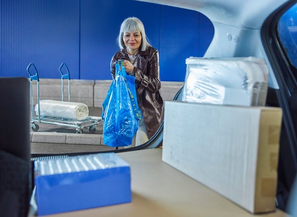 A smiling person loading a blue FRAKTA shopping bag and boxed items into the boot of their car. A rolled-up mattress can be seen in the background on a trolley.