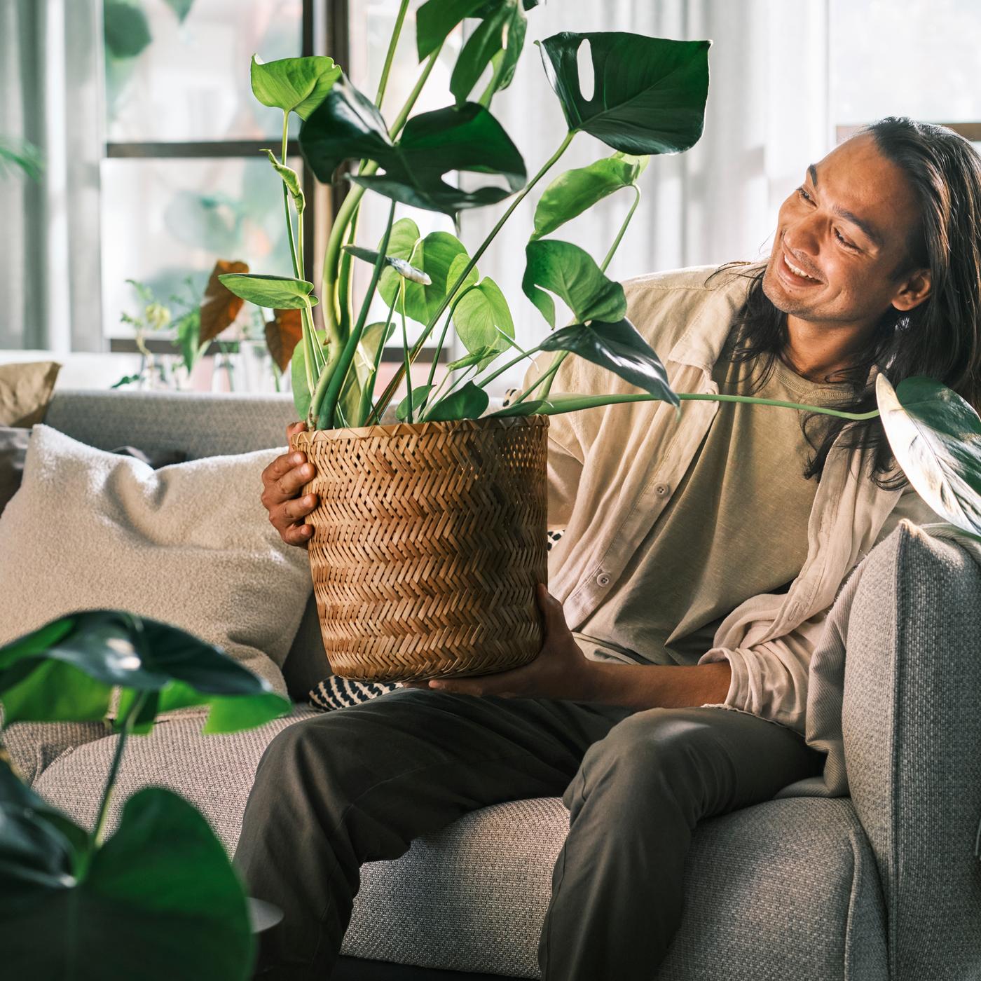 A smiling person sitting on a light-grey ÄPPLARYD three-seat sofa holding a monstera planted in a KLYNNON bamboo plant pot.